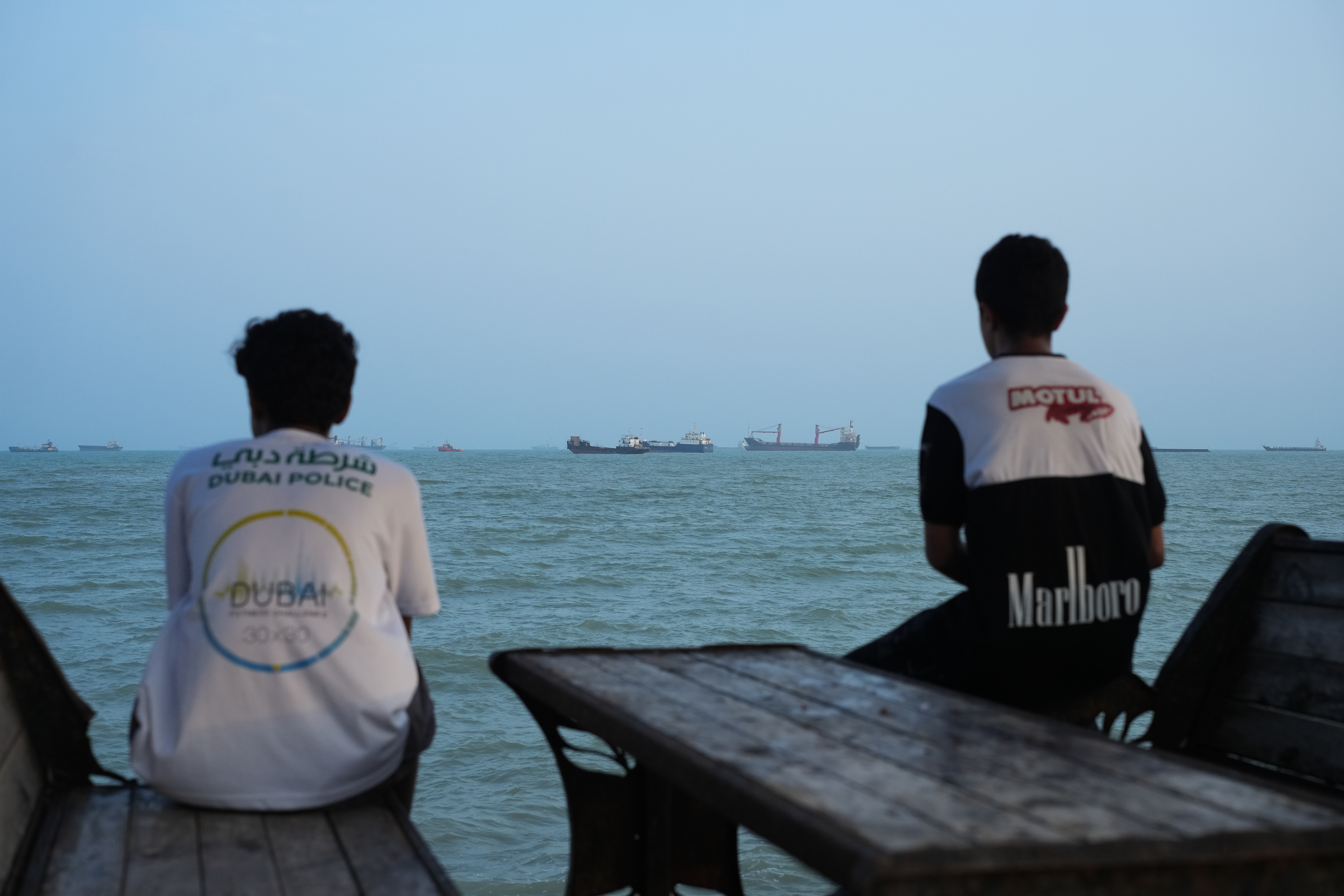 Two youth sit on the shoreline as ships are seen anchored in the Strait of Hormuz on August 10, 2026 off the coast of Bandar Abbas, Iran