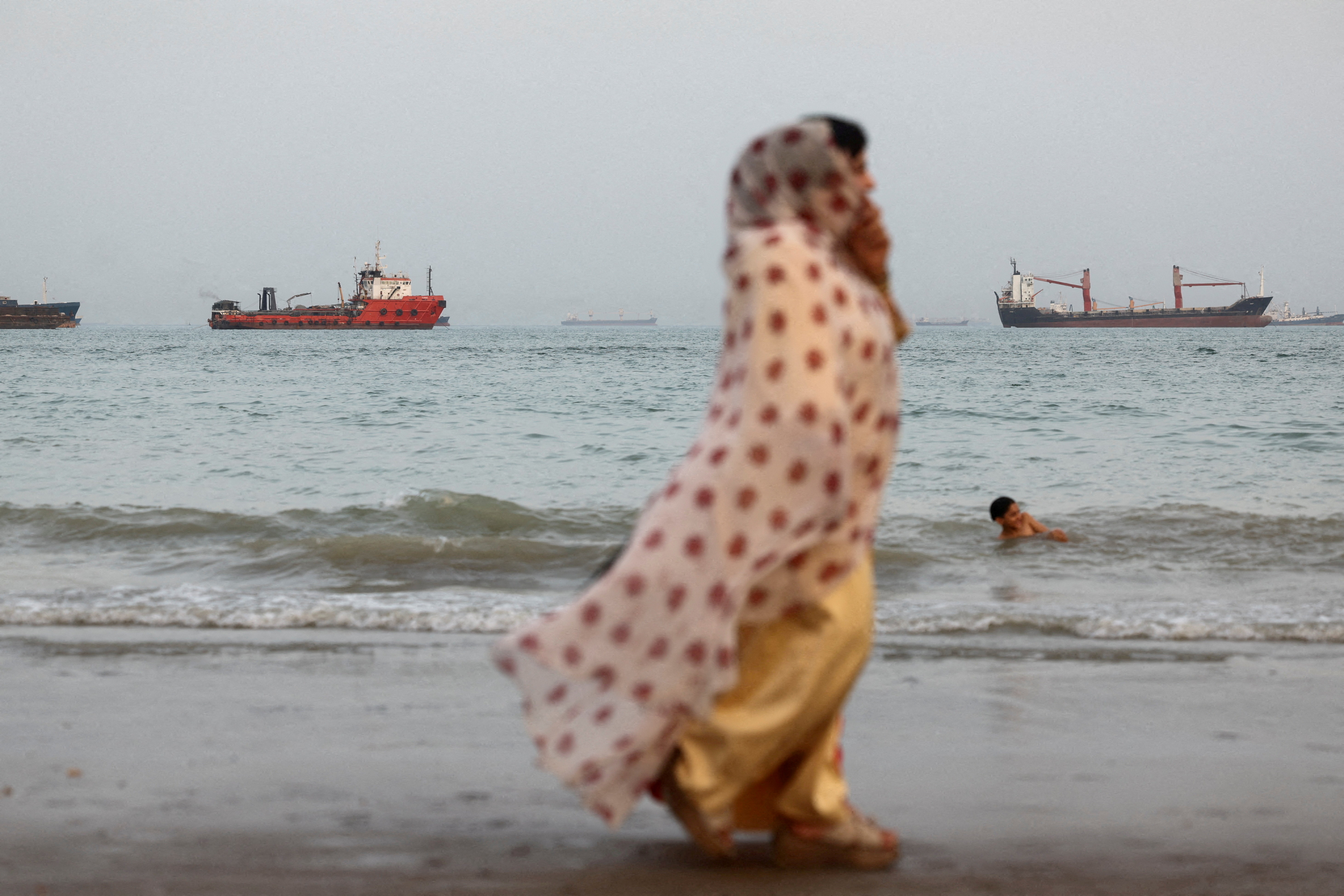 People walk on the beach, with vessels in the sea behind them.