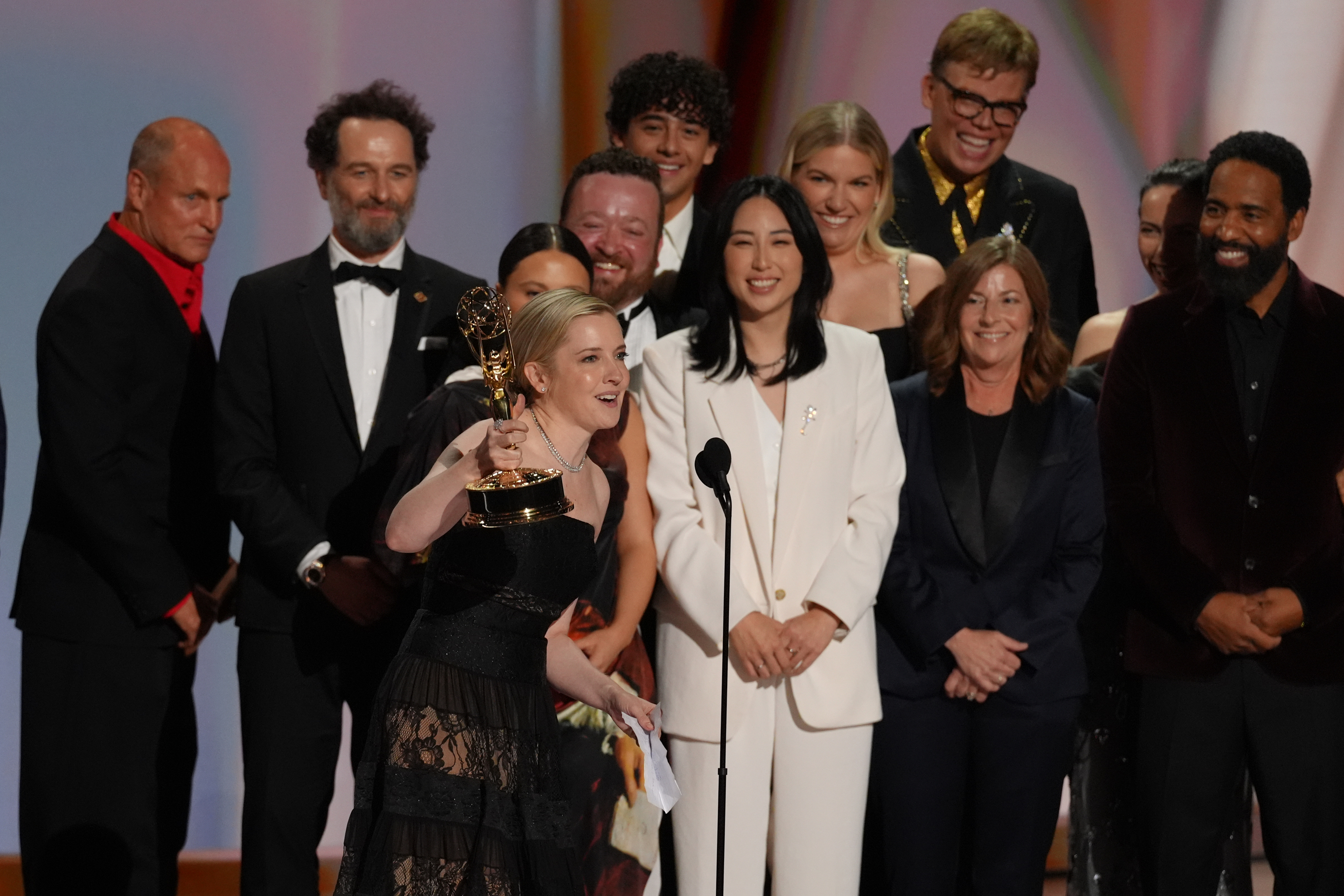 A woman stands at a lectern holding an Emmy statuette with a group of people behind her.