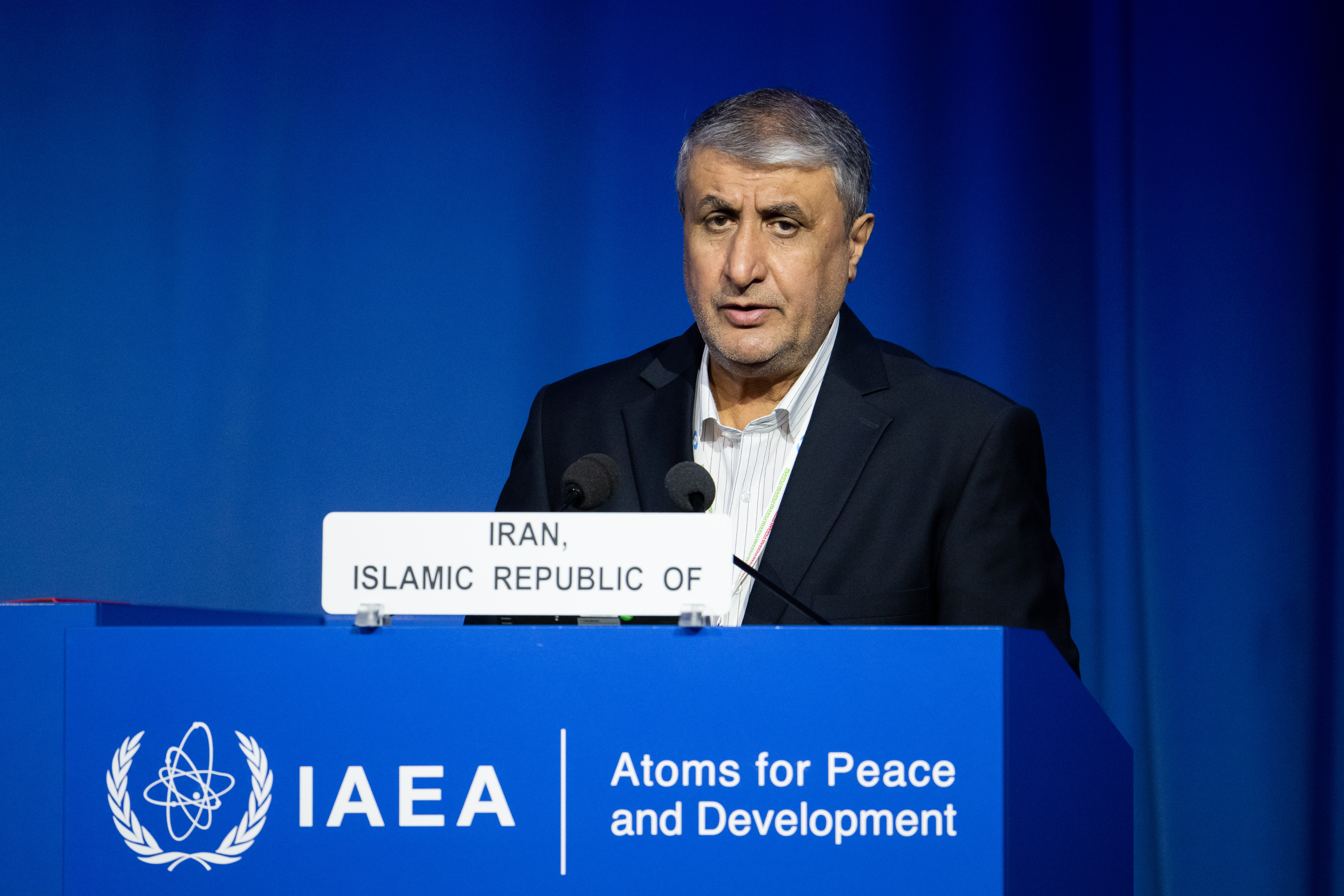 A man in a dark suit speaks into a microphone at a lectern in a conference hall.