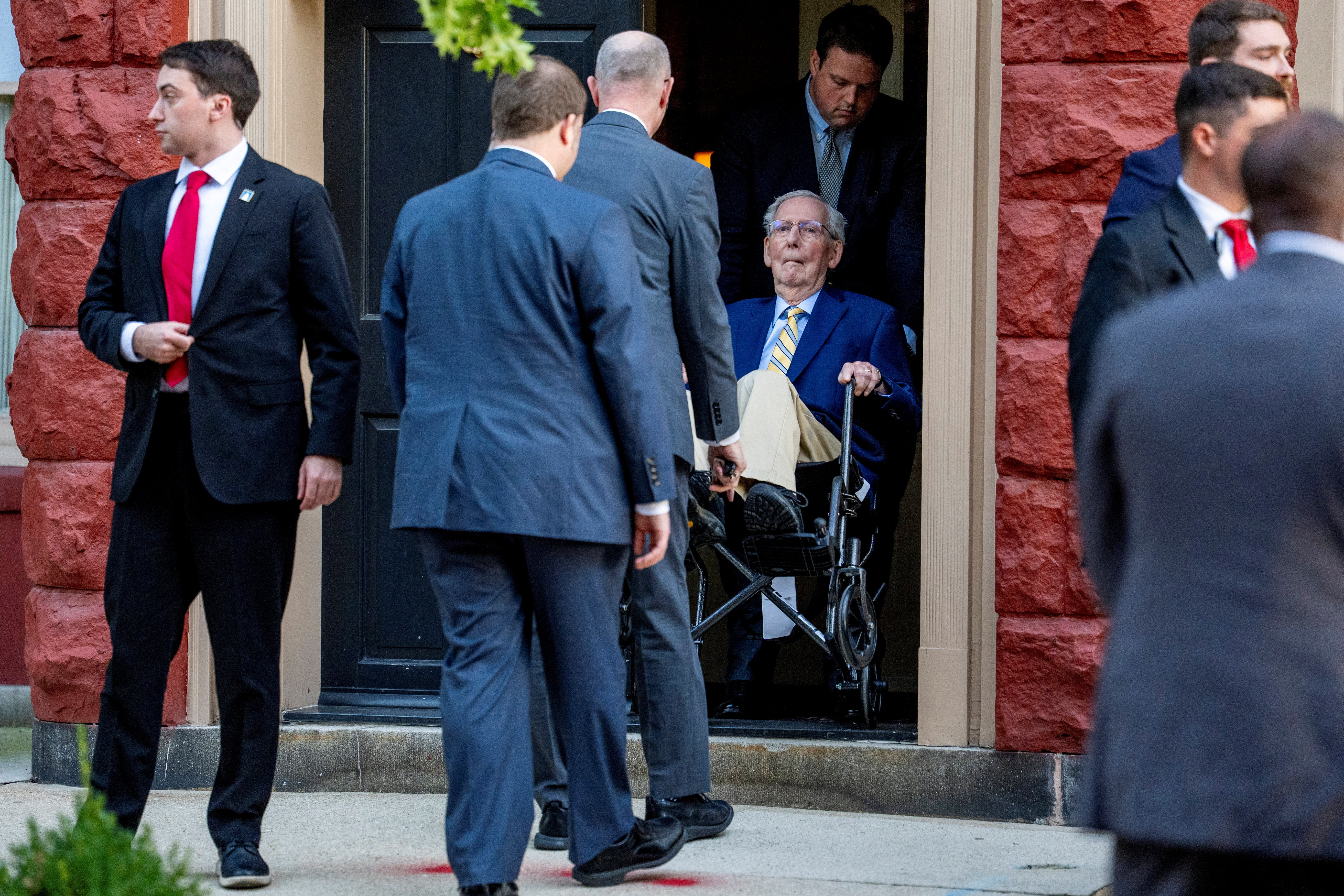 Senator Mitch McConnell sits in a wheelchair with other men in suits standing around.