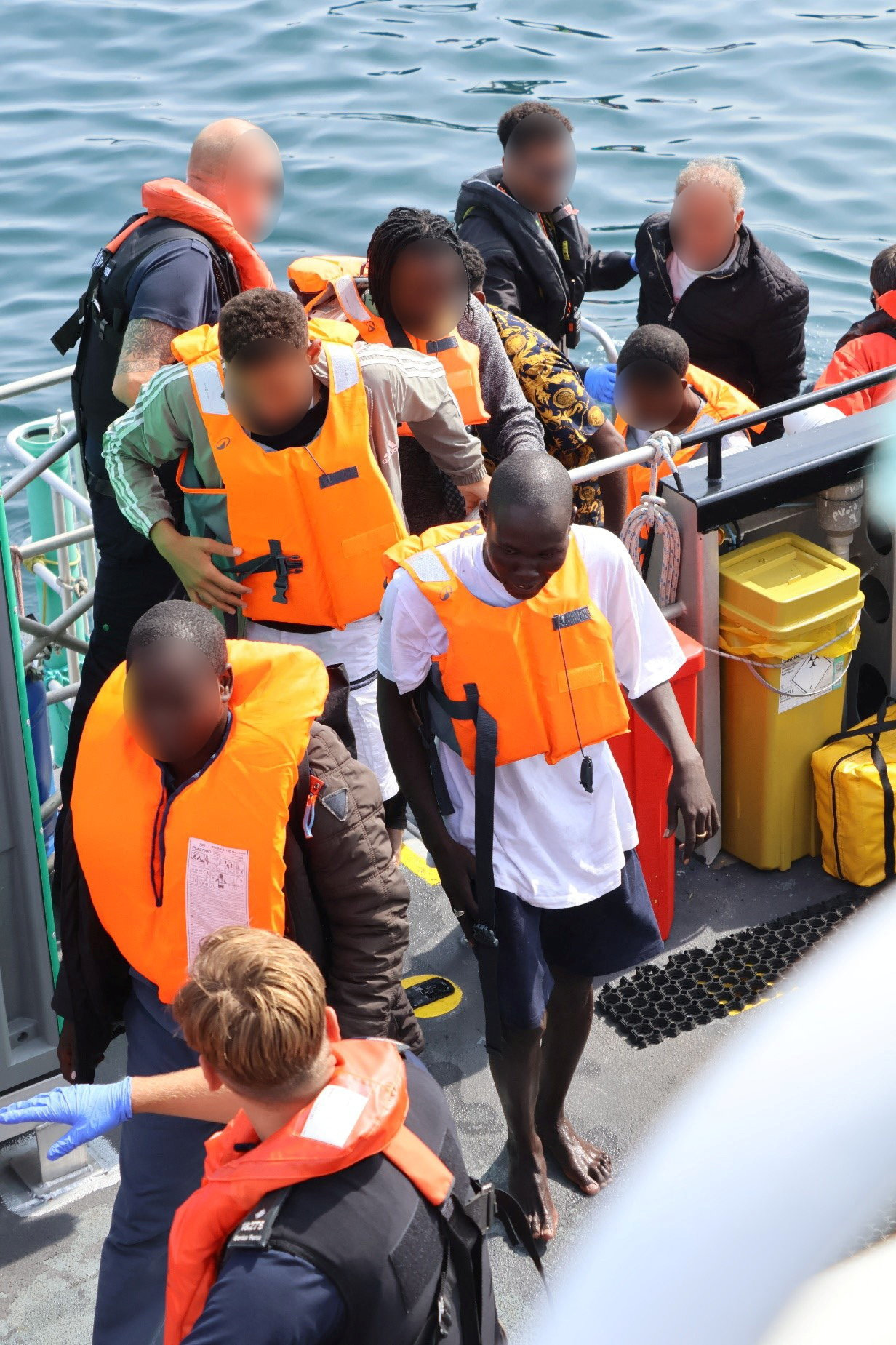 An overcrowded inflatable boat packed with people, their faces blurred, seen from above on open water.